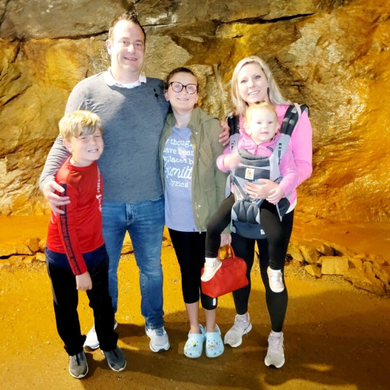 A family including a husband and wife, preteen girl, school-aged boy, and toddler girl in a baby carrier pose in front of rock walls in the Consolidated Gold Mine.