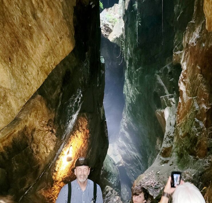 A former gold miner dressed in a black and white plaid shirt and black suspenders with a black hard hat on leads a group of people through towering rock walls and caverns.