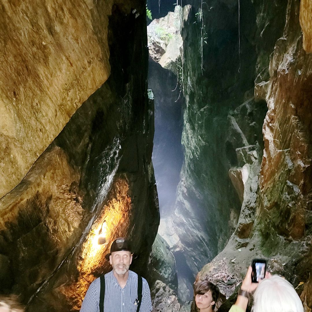 A former gold miner dressed in a black and white plaid shirt and black suspenders with a black hard hat on leads a group of people through towering rock walls and caverns.