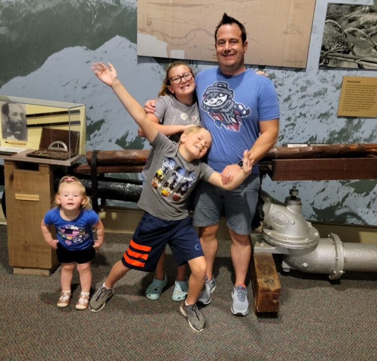 A dad in a blue Trash Pandas shirt poses with his three children in front of a gold mining exhibit at the Dahlonega Gold Museum.