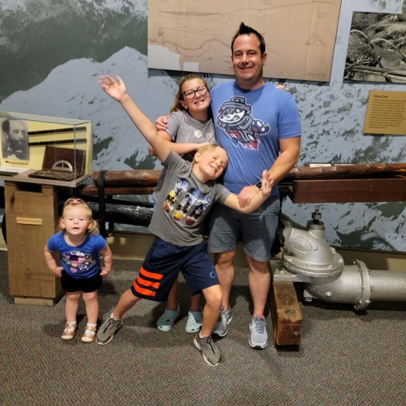 A dad in a blue Trash Pandas shirt poses with his three children in front of a gold mining exhibit at the Dahlonega Gold Museum.