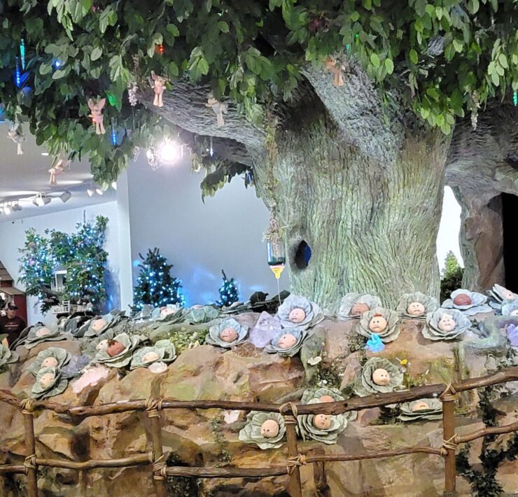 A large tree sits with a patch of cabbages beneath it with cabbage patch doll heads coming from the cabbages.
