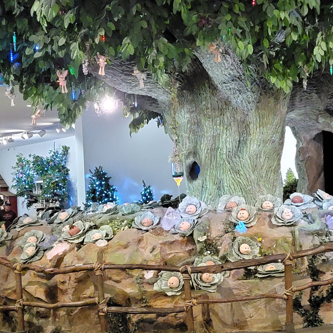 A large tree sits with a patch of cabbages beneath it with cabbage patch doll heads coming from the cabbages.