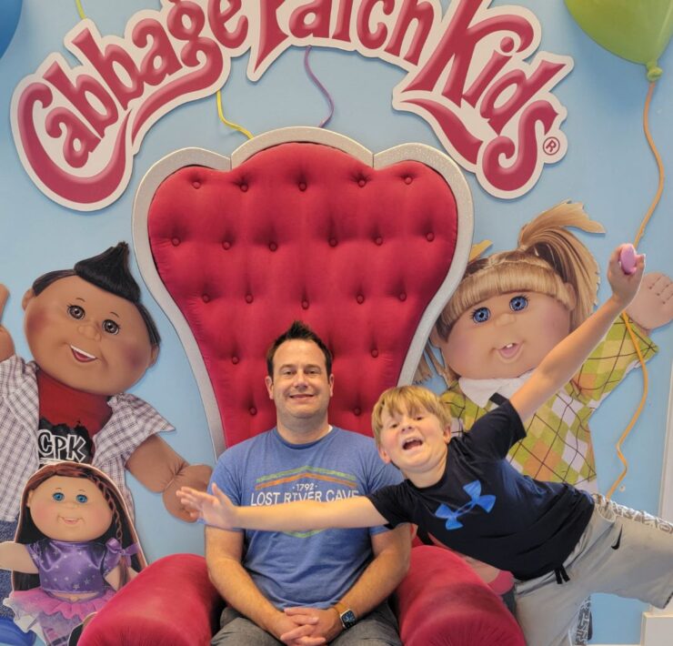 A man sits in a red throne-like chair with a school-aged boy leaning into the frame with the words "Cabbage Patch Kids" behind them.