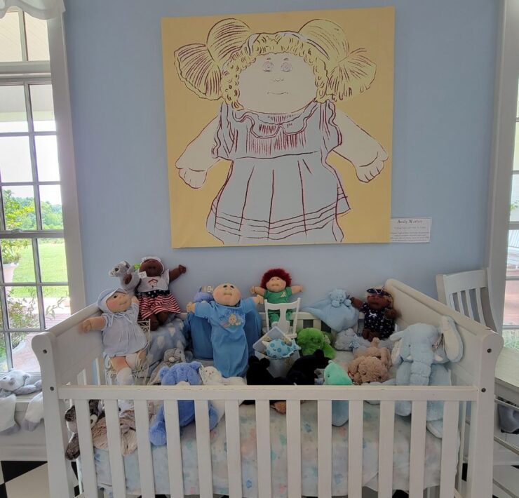 A group of Cabbage Patch Kid dolls and stuffed animals fill a white crib in front of a blue wall and Cabbage Patch Doll painting in Dahlonega, GA.