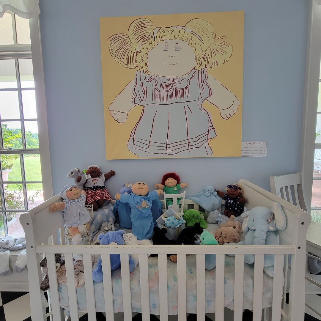 A group of Cabbage Patch Kid dolls and stuffed animals fill a white crib in front of a blue wall and Cabbage Patch Doll painting in Dahlonega, GA.