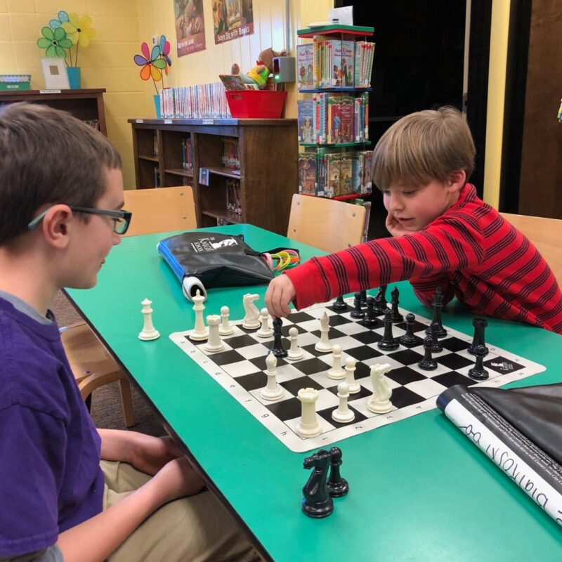 A young boy in a red striped shirt leans across a green table to move his chess piece at the Knight School Chess Academy.