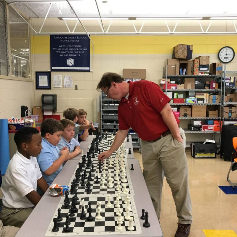 An instructor in a red shirt plays against a row of students behind their own chess boards.