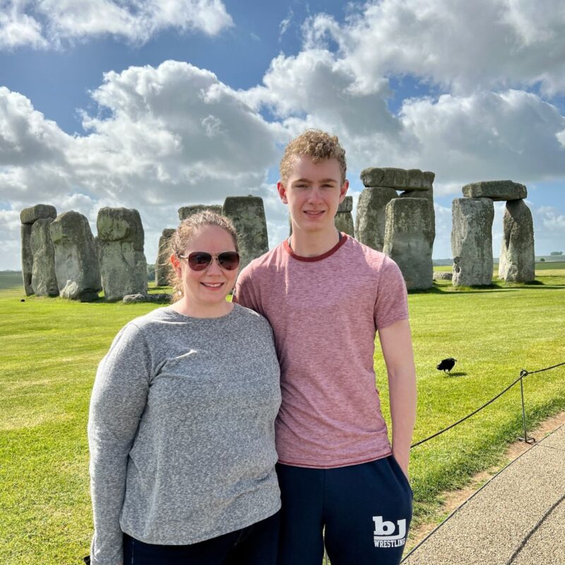 Awesome Mom, Melanie stands with one of her sons in front of a circle of standing stones while traveling abroad. 