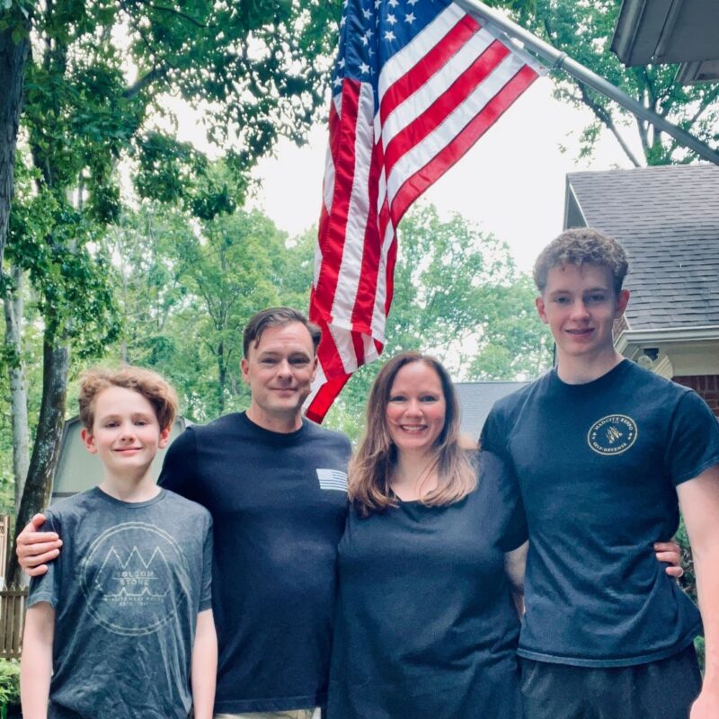 A family including Awesome Mom, Melanie, stands in front of an American flag at the entrance of their home. 