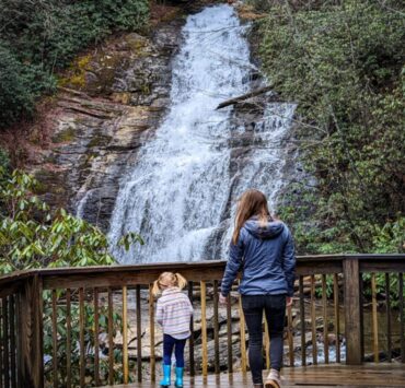 A young girl in bright blue rain boots and a rain jacket walks alongside a slightly older girl in a gray rain jacket on an observatory deck of a waterfall in the North Georgia mountains.
