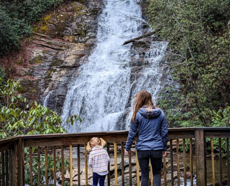 A young girl in bright blue rain boots and a rain jacket walks alongside a slightly older girl in a gray rain jacket on an observatory deck of a waterfall in the North Georgia mountains.