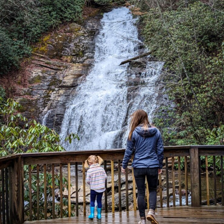 A young girl in bright blue rain boots and a rain jacket walks alongside a slightly older girl in a gray rain jacket on an observatory deck of a waterfall in the North Georgia mountains.