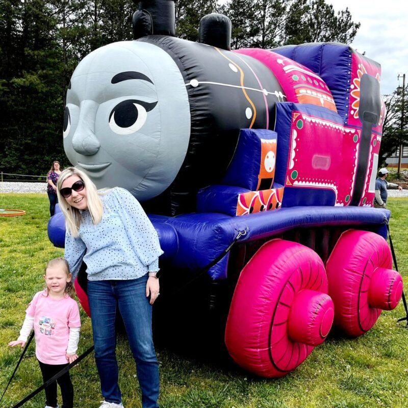 A toddler girl and pink and her mother stand in front of a pink Thomas the Train character.