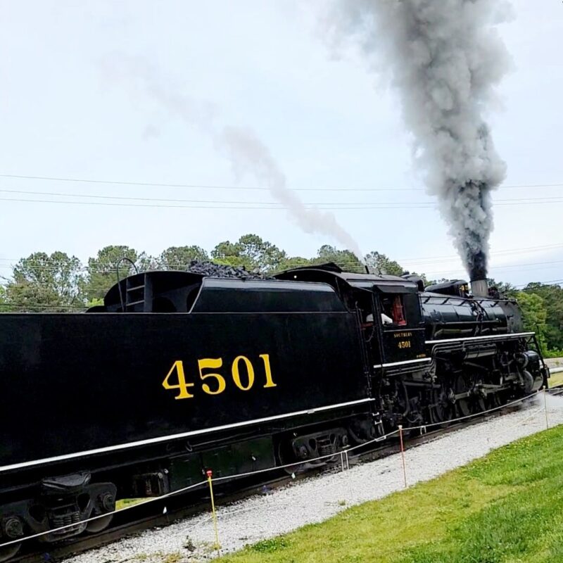 A black train engine with smoke billowing from it makes its way down the railroad tracks at the Tennessee Valley Railroad Museum.